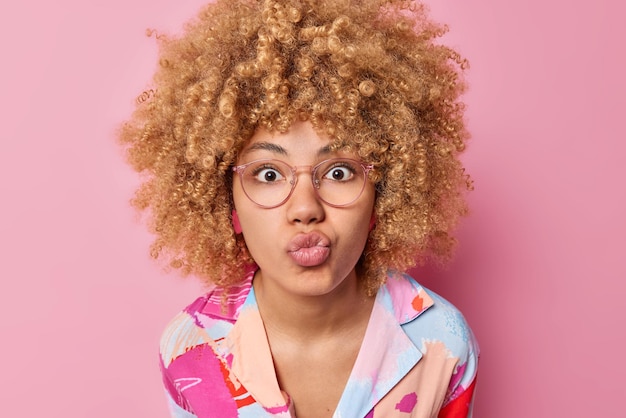 Foto mensen en gezichtsuitdrukkingen concept afgevraagd krullend haired vrouw kijkt verrassend naar camera houdt lippen gevouwen draagt transparante bril en kleurrijk shirt geïsoleerd over roze achtergrond