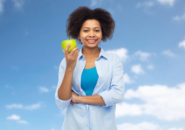 mensen, eten, gezond eten en tandheelkundige zorg concept - gelukkige Afro-Amerikaanse jonge vrouw met groene appel over blauwe lucht en wolken achtergrond