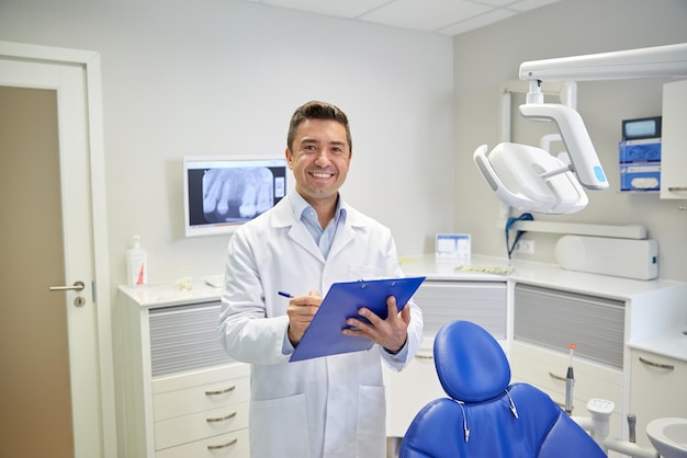 mensen, geneeskunde, stomatologie en gezondheidszorgconcept - gelukkige mannelijke tandarts van middelbare leeftijd in witte jas die naar klembord schrijft op het kantoor van de tandheelkundige kliniek