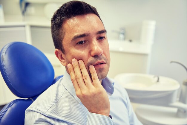 Foto mensen, geneeskunde, stomatologie en gezondheidszorgconcept - ongelukkige mannelijke patiënt met kiespijn zittend op een tandartsstoel op het kantoor van de kliniek