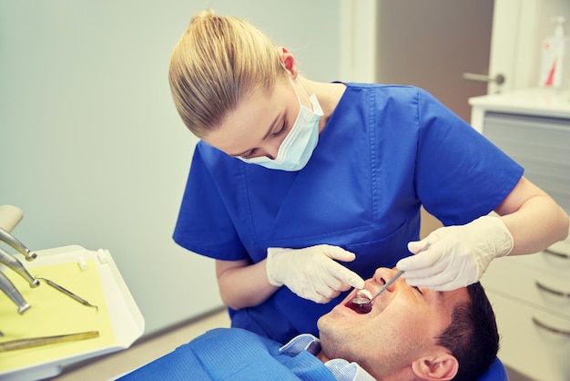 Foto mensen, geneeskunde, stomatologie en gezondheidszorgconcept - vrouwelijke tandarts met tandspiegel die de tanden van de mannelijke patiënt controleert op het kantoor van de tandheelkundige kliniek