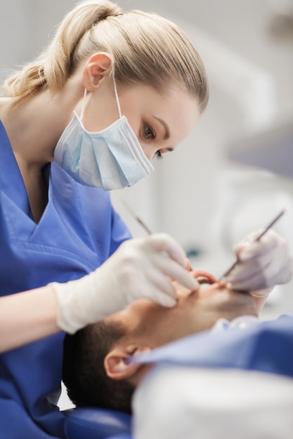 Foto mensen, geneeskunde, stomatologie en gezondheidszorgconcept - vrouwelijke tandarts met tandspiegel en sonde die de tanden van de mannelijke patiënt controleert op het kantoor van de tandheelkundige kliniek