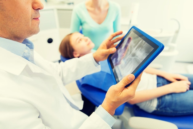 mensen, geneeskunde, stomatologie, technologie en gezondheidszorgconcept - close-up van mannelijke tandarts met röntgenfoto op het computerscherm van tablet-pc en geduldig meisje met haar moeder op het kantoor van de tandheelkundige kliniek