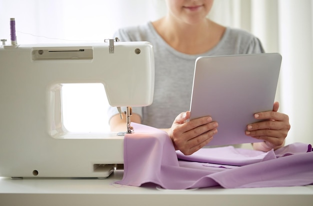 mensen, handwerk, technologie en maatwerkconcept - kleermaker vrouw met naaimachine, tablet pc en stof