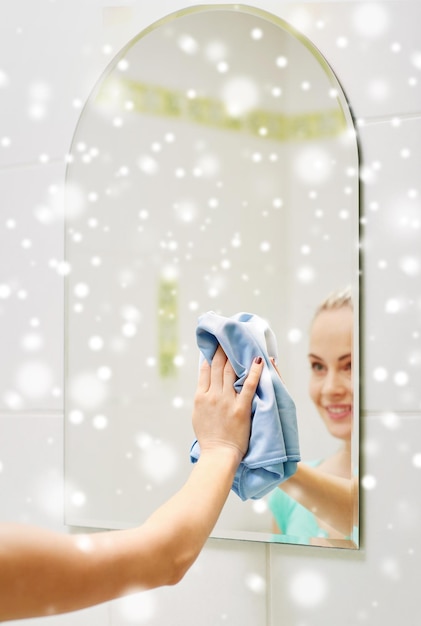mensen, huishoudelijk werk en huishoudconcept - close-up van gelukkige vrouw die spiegel met doek thuis schoonmaakt over sneeuweffect