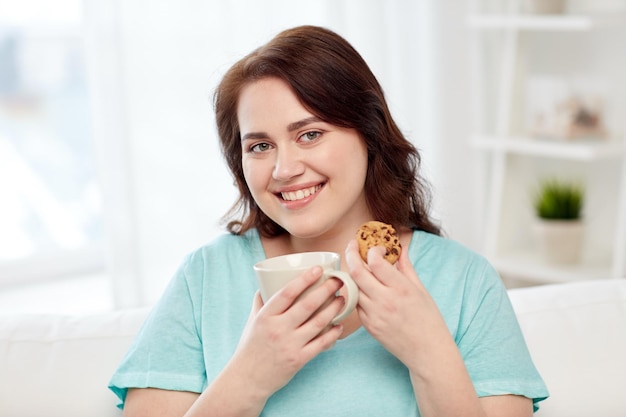 mensen, junkfood, ongezond eten en vrije tijd concept - gelukkige plus size jonge vrouw met kopje thee met koekje thuis