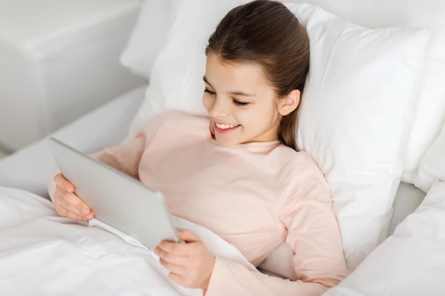 mensen, kinderen, rust en technologie concept - gelukkig lachend meisje wakker liggen met tablet pc-computer in bed thuis