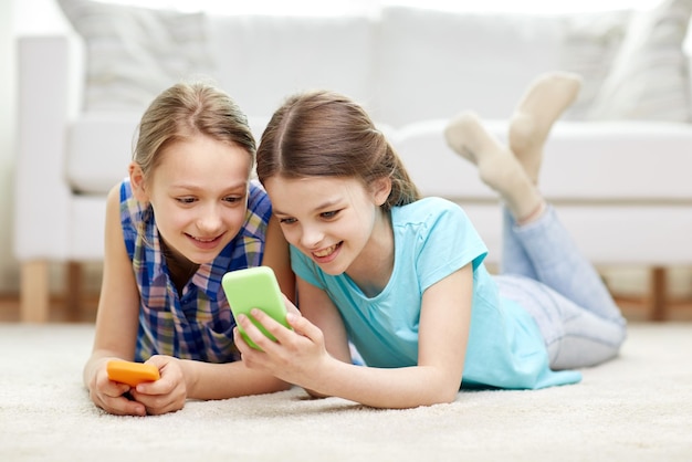 Mensen, kinderen, technologie, vrienden en vriendschapsconcept - gelukkige kleine meisjes met smartphones die thuis op vloer liggen