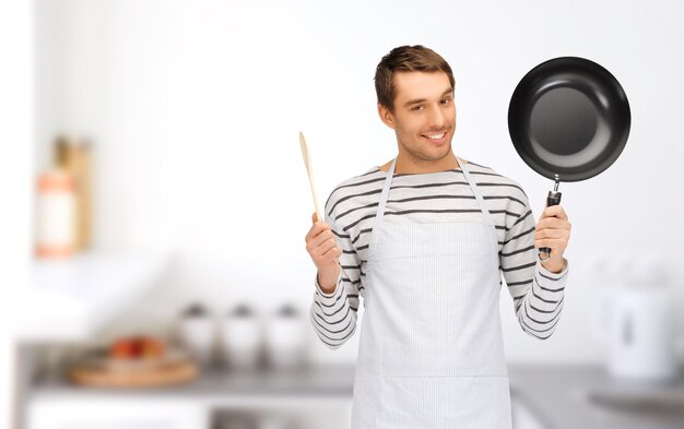 mensen, koken en culinair concept - gelukkige man of kok in schort met koekenpan en houten lepel over de achtergrond van de huiskeuken