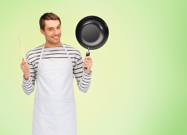 mensen, koken en culinair concept - gelukkige man of kok in schort met koekenpan en houten lepel over groene natuurlijke achtergrond