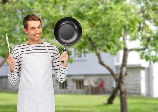 Foto mensen, koken en culinair concept - gelukkige man of kok in schort met koekenpan en houten lepel over zomertuin en huisachtergrond