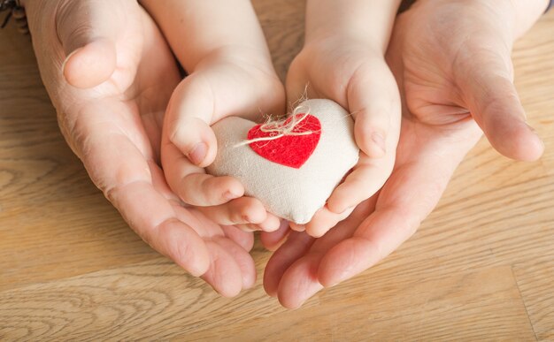 Mensen, leeftijd, familie, liefde en gezondheidszorgconcept - sluit omhoog van hogere vrouw en weinig jongenshanden houdend rood hart over houten achtergrond