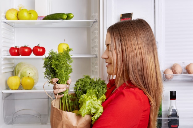 Mensen, levensstijl en gezond eten concept. zijwaarts schot van schattige vrouw houdt papieren doos met dille en sla, heeft een koelkast vol met groenten en fruit, gaat vegetarische verse salade maken