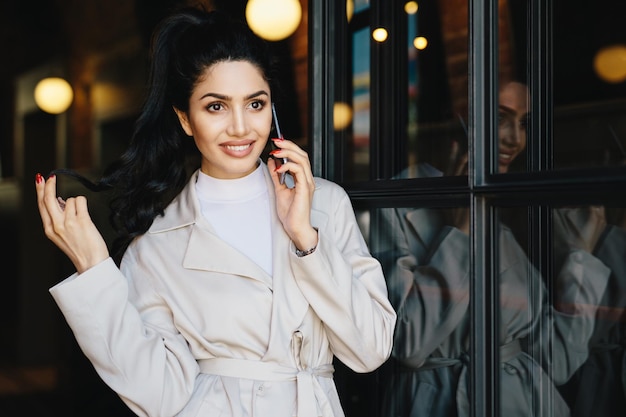 Mensen levensstijl moderne technologieën en mode concept portret van modieuze vrouw met donker haar heldere ogen met lange wimpers en volle lippen dragen witte jas met smartphone buitenshuis