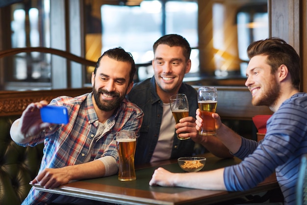 mensen, mannen, vrije tijd, vriendschap en technologieconcept - gelukkige mannelijke vrienden die bier drinken en selfie nemen met smartphone in bar of pub