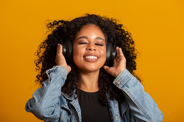 Mensen, muziek, emoties concept. Opgetogen zorgeloze vrouw met Afro kapsel danst in ritme van melodie, sluit ogen luistert luid lied in koptelefoon. Mooie afro vrouw met haar koptelefoon