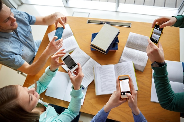 mensen, onderwijs, technologie en examenconcept - close-up van studenten met smartphones die een foto van de boekenpagina maken en spiekbriefjes maken in de schoolbibliotheek