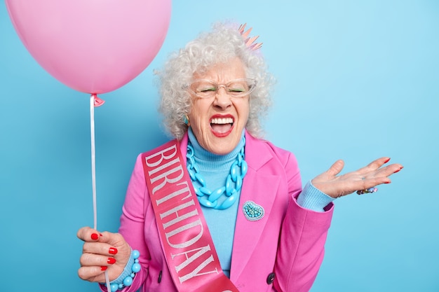 Mensen oude mensen vakantie concept. Emotionele senior vrouw roept luid en drukt negatieve emoties uit houdt mond open, gekleed in feestelijke kleding houdt opgeblazen ballon vast