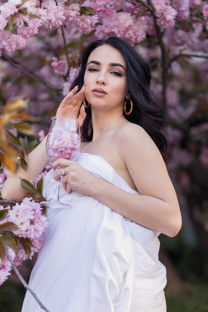 Mensen, rust, comfort en vrije tijd concept - Jonge vrouw door de avondstraten gewikkeld in een deken. Mooi jong meisje in het park en een bloeiende boomsakura met een glas met bloemblaadjes