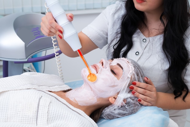 Mensen, schoonheid, spa, cosmetologie en technologie concept - close-up van mooie jonge vrouw liggend met gesloten ogen met gezichtsmassage door massager in spa.