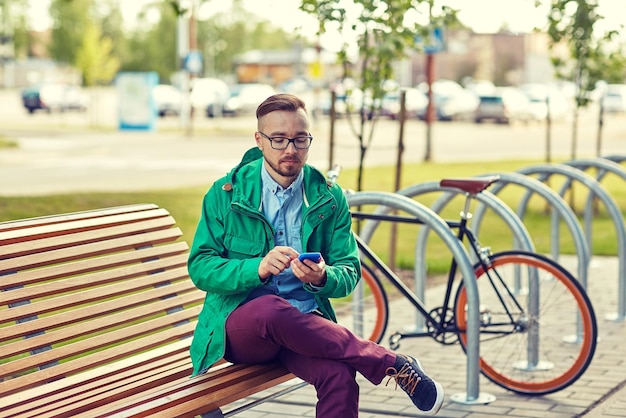 mensen, stijl, technologie en levensstijl - gelukkige jonge hipster man met smartphone en fixie fiets zittend op een bankje in de stad