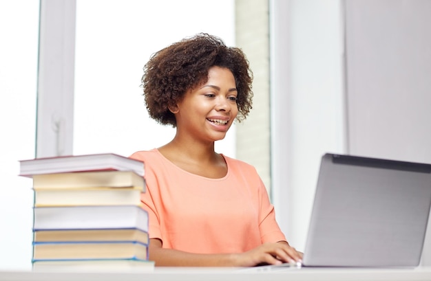 mensen, technologie en onderwijsconcept - gelukkige Afro-Amerikaanse jonge vrouw die aan tafel zit met een laptopcomputer en boeken thuis