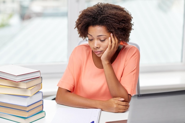 mensen, technologie en onderwijsconcept - verveelde Afro-Amerikaanse jonge vrouw zittend aan tafel met laptopcomputer, boeken en notitieblok thuis