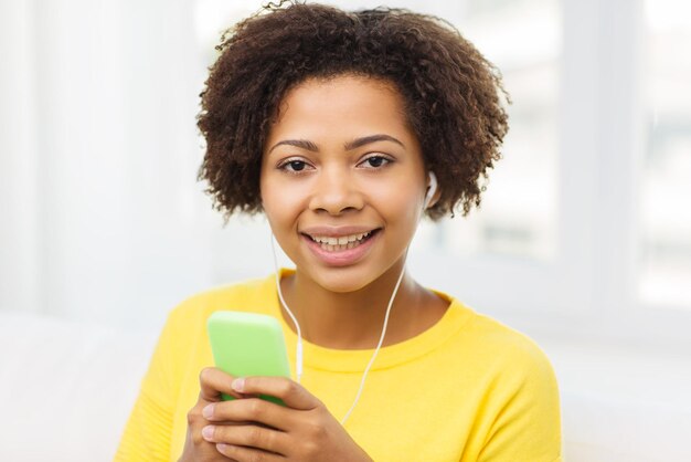 mensen, technologie en vrije tijd concept - gelukkige Afro-Amerikaanse jonge vrouw zittend op de bank met smartphone en koptelefoon luisteren naar muziek thuis