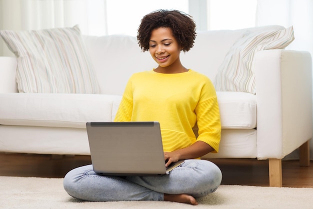 mensen, technologie en vrije tijd concept - gelukkige Afro-Amerikaanse jonge vrouw zittend op de vloer met laptopcomputer thuis