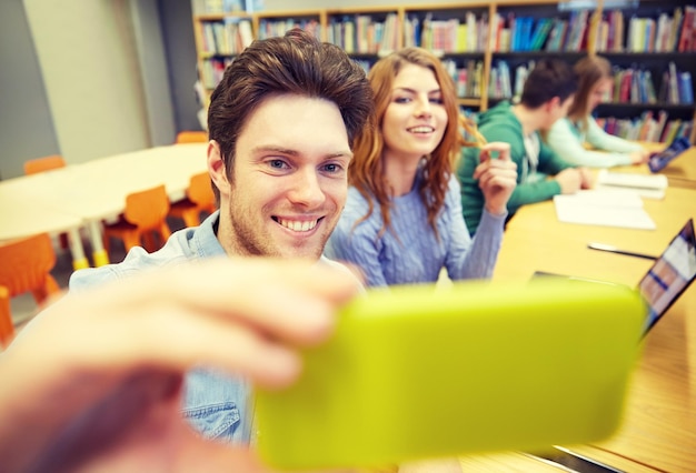 Foto mensen, technologie, onderwijs en schoolconcept - gelukkige studenten of vrienden met smartphone die selfie in bibliotheek nemen