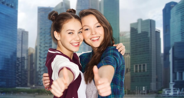 mensen, tieners, vriendschap, reizen en toerisme concept concept - gelukkig lachende mooie tienermeisjes knuffelen en duimen opdagen over singapore stad achtergrond