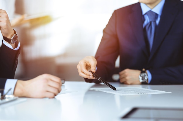 Mensen uit het bedrijfsleven bespreken een contract tijdens vergadering of onderhandeling, zitten samen aan de balie en een onbekende vrouw op de achtergrond, close-up. Groep onbekende zakenlieden in een zonnig modern kantoor.