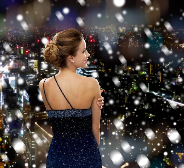 mensen, vakantie, kerstmis en mensenconcept - glimlachende vrouw in avondjurk over besneeuwde nachtstadsachtergrond van achteren