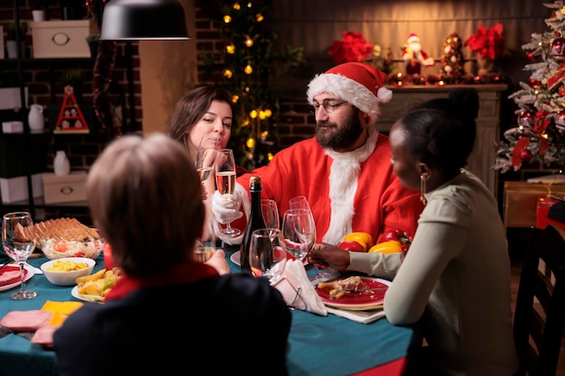 Mensen vieren kerstmis, rammelende mousserende wijnglazen aan feestelijke tafel, man in kerstmankostuum die toast voorstelt. Wintervakantieviering, samenzijn met grote familie