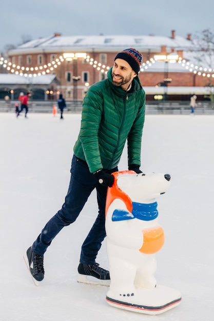 Mensen vrije tijd actieve levensstijl concept Vrolijke man van middelbare leeftijd leert schaatsen gebruikt skatehulp besteedt vrije tijd op de ijsbaan kijkt vreugdevol opzij geniet ervan om weekenden actief door te brengen