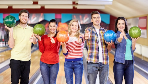 mensen, vrije tijd, sport, vriendschap en entertainment concept - gelukkige vrienden die ballen vasthouden en duimen opdagen in de bowlingclub