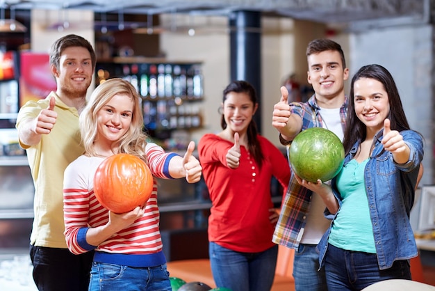 mensen, vrije tijd, sport, vriendschap en entertainment concept - gelukkige vrienden die ballen vasthouden en duimen opdagen in de bowlingclub