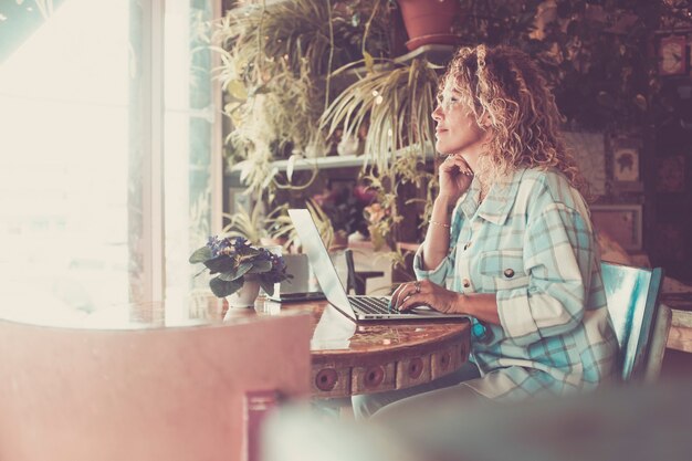 Mensen werken thuis in een slimme werkbaan met een laptopcomputer die op tafel zit en buiten het raam kijkt - gelukkige volwassen vrouw glimlacht en geniet van online moderne levensstijl