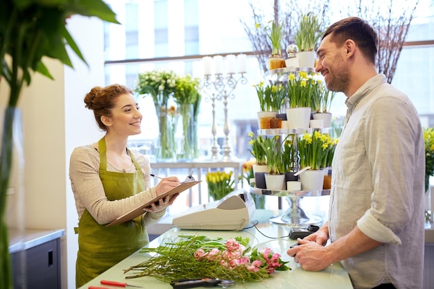 mensen, winkelen, verkoop, bloemisterij en consumentisme concept - gelukkige lachende bloemist vrouw met klembord en man of klant die bestelling plaatst bij bloemenwinkel