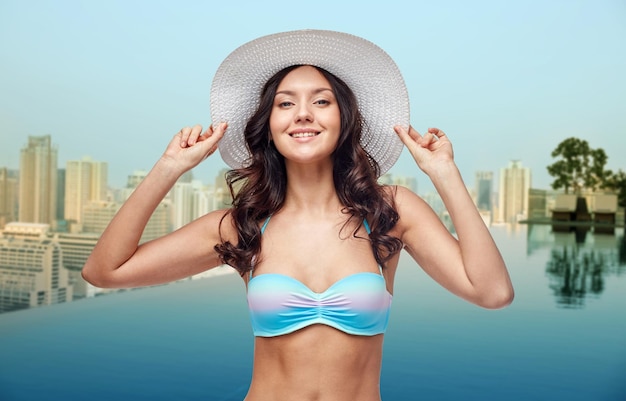 mensen, zomervakantie, reizen en toerisme concept - gelukkige jonge vrouw in bikini badpak en zonnehoed over oneindigheid rand zwembad en stad achtergrond