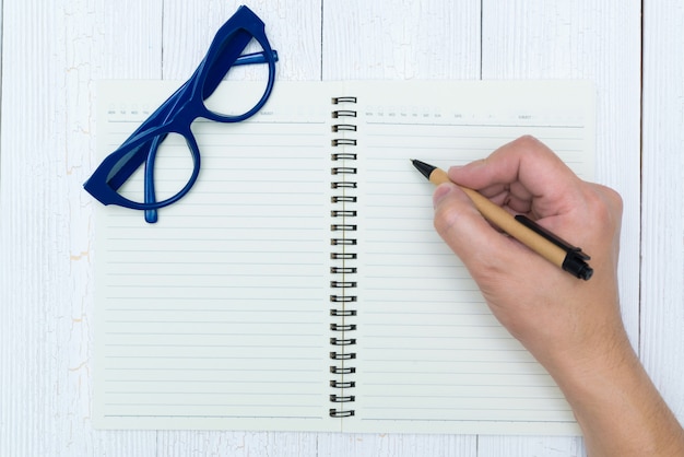 Mensenhand die op blanco pagina van notitieboekjedocument schrijven met pen, glazen en bureaulevering, hoogste mening.