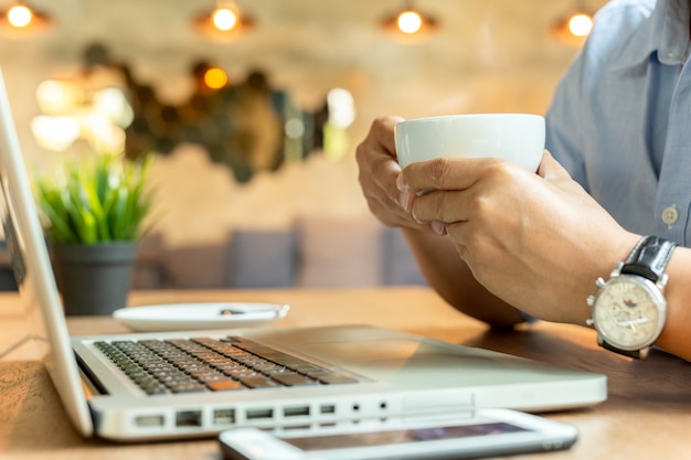 Mensenhanden die kopkoffie met mobiele telefoon en laptop op houten lijst houden.