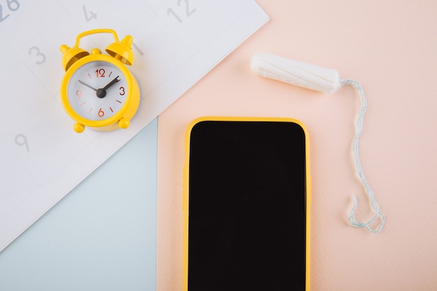 Menstrual cycle concept. Yellow alarm clock and a mobile application on the smartphone screen. Cotton tampon on the background.