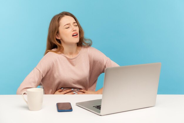 Menstruatiekrampen, buikpijn op het werk. Vrouwelijke werknemer die een pijnlijke buik vasthoudt, ongemak voelt, last heeft van brandend maagzuur, gastro-intestinale stoornis. indoor studio-opname geïsoleerd op blauwe achtergrond