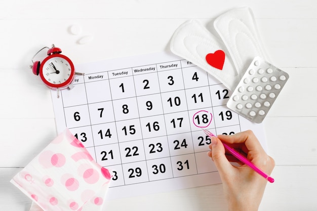 Menstruation calendar with pads, alarm clock, hormonal contraceptive pills. Female's menstrual cycle concept. Pain relievers for menstrual pain