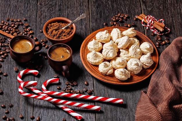 머랭 쿠키는 어두운 나무 배경에 크리스마스 사탕수수, 커피 컵, 커피 콩, 갈색 설탕, 계피 스틱이 있는 접시에 제공됩니다.