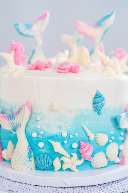 Mermaid-themed 3 layer vanilla cake decorated with chocolate mermaid tails and seashells on a white cake stand.