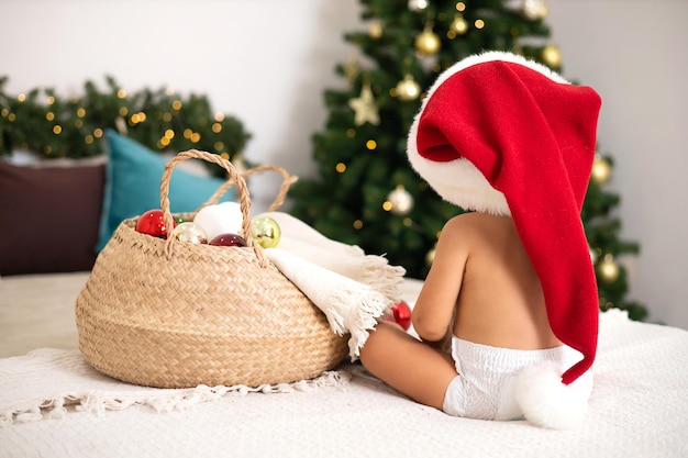 사진 메리 크리스마스 휴일 크리스마스 트리 라이프스타일을 위해 장식된 아늑한 침실에 기저귀와 산타39의 모자를 쓴 아이가 침대에 앉아 있습니다.