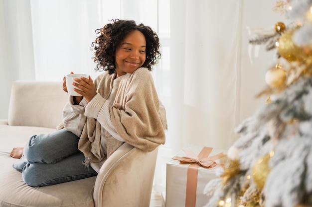 Merry christmas african american woman with cup of hot drink coffee tea sitting near christmas tree