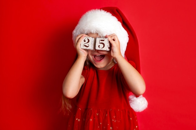 메리 크리스마스와 해피 홀리데이 산타 모자를 쓴 재미있는 소녀가 숫자 25가 있는 큐브를 들고 있습니다